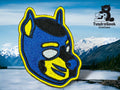 Close-up of a custom embroidered pup hood patch in blue and yellow with metallic thread accents, shown against a mountain and lake backdrop.