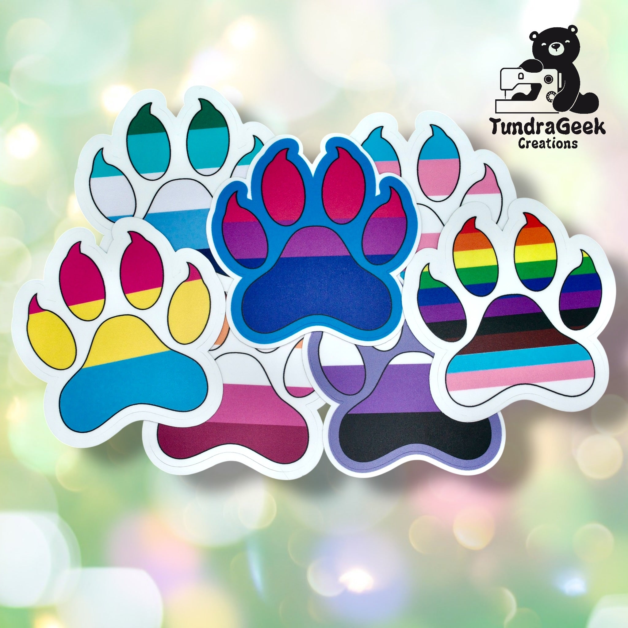 A collection of paw print stickers featuring various LGBTQ+ pride flag colors, arranged on a blurred pastel background with the TundraGeek Creations logo in the corner.