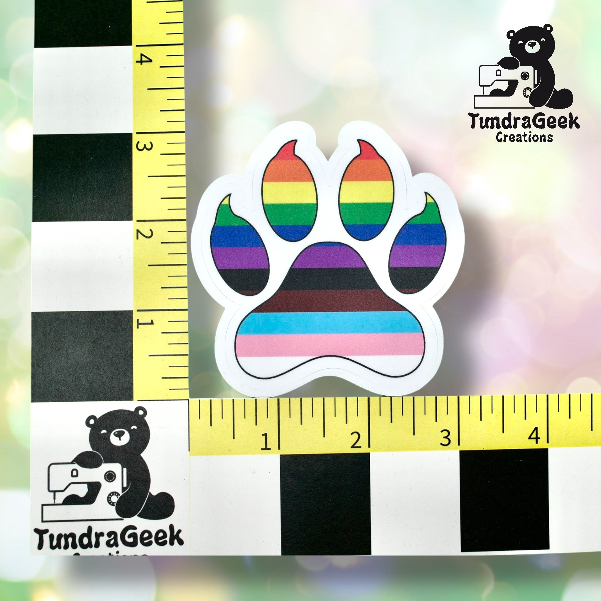 Sticker of a paw print with rainbow and LGBTQ+ flag colors placed between two rulers for scale, showing a size of about 2.5 inches.