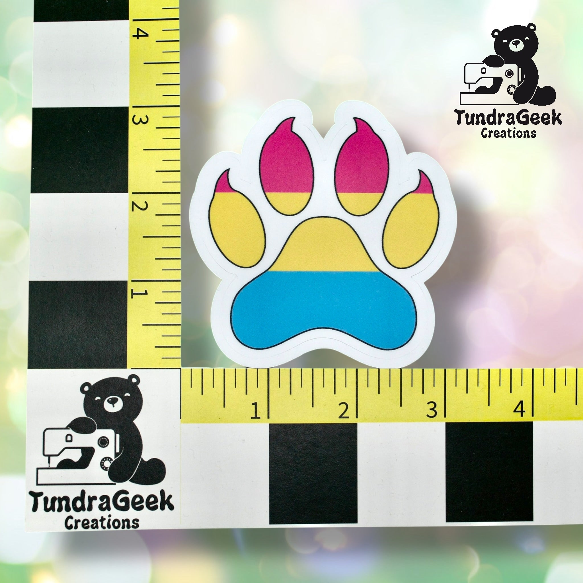 A sticker of a paw print with pink, yellow, and blue sections is placed next to a ruler on a checkered background. TundraGeek Creations logo appears twice.