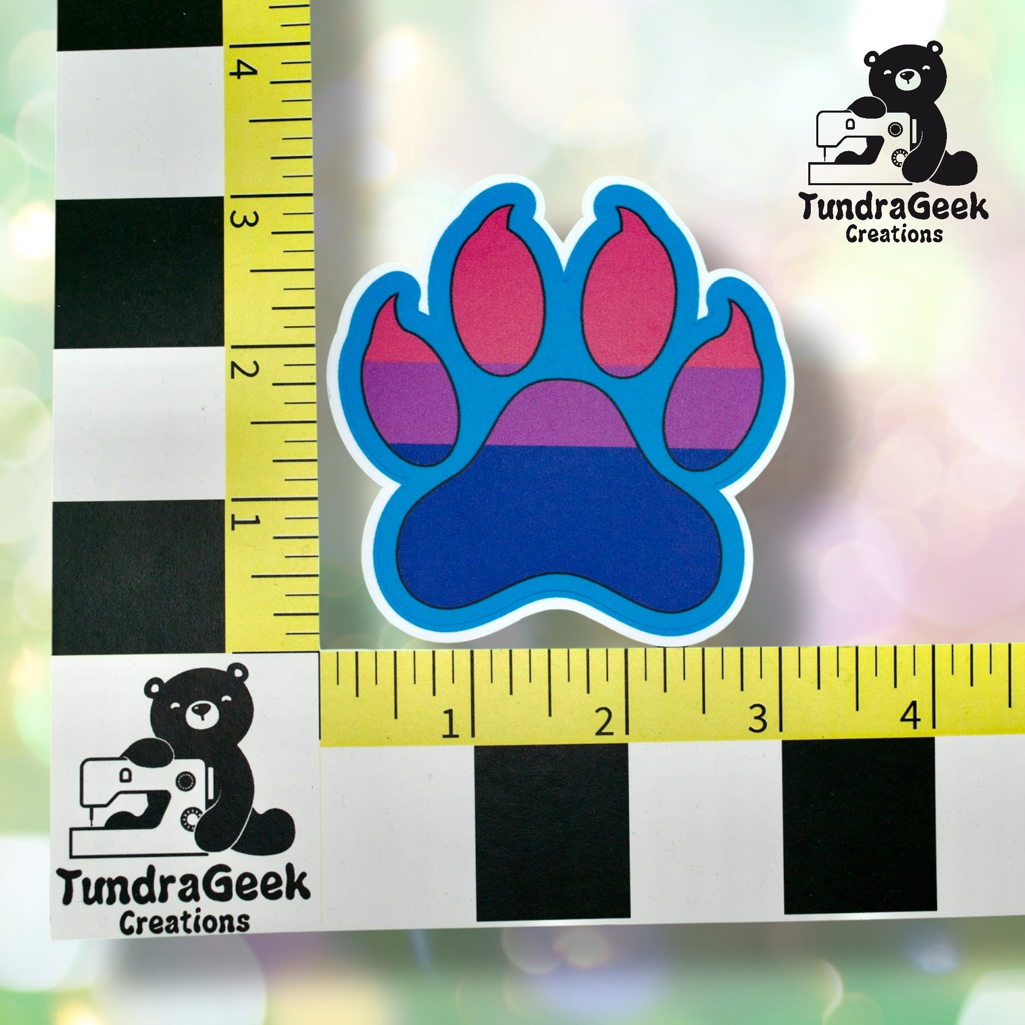 A sticker of a paw print with pink, purple, and blue stripes is measured by a ruler grid with TundraGeek Creations branding in the corners.