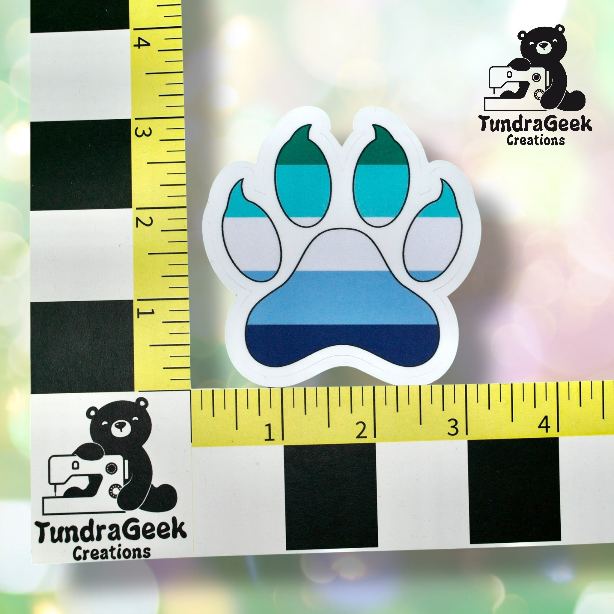 A sticker shaped like a paw print with stripes in shades of blue and teal is placed next to a ruler with TundraGeek Creations branding for scale.