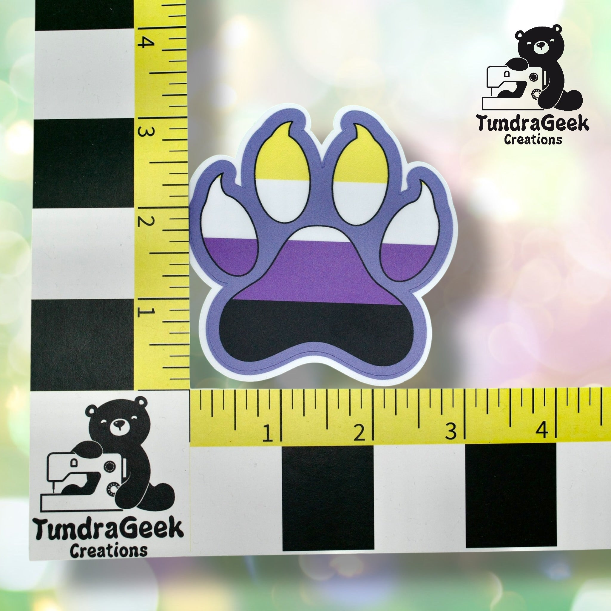 A paw print sticker featuring the nonbinary pride flag colors is placed between two measuring rulers, with a TundraGeek Creations logo in the corners.