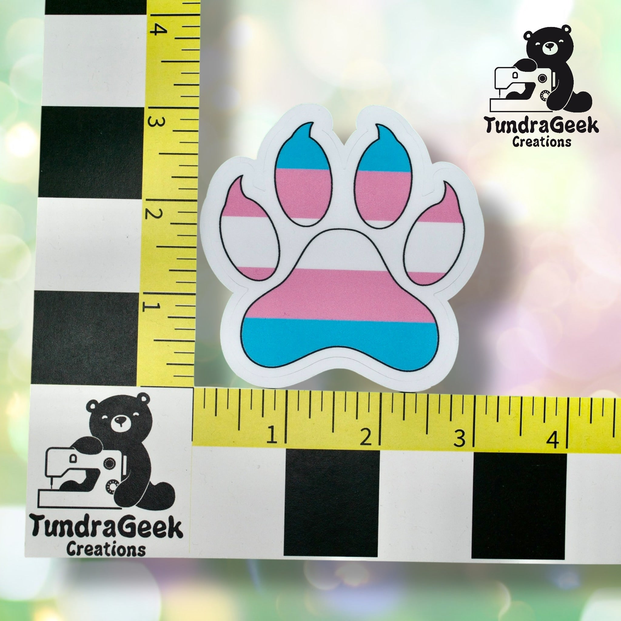 A sticker shaped like a paw print with the transgender pride flag colors, placed on a measuring grid. The TundraGeek Creations logo appears in two corners.