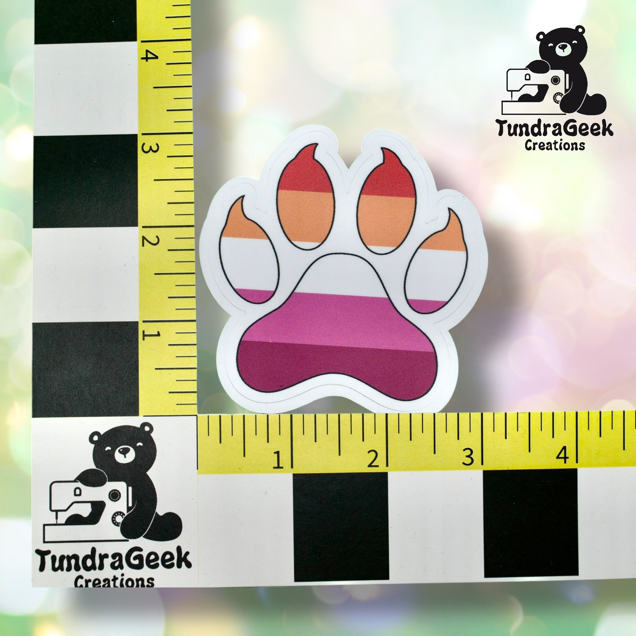 A sticker shaped like a paw print with shades of orange, red, and pink sits on a measuring grid with a TundraGeek Creations logo in the corners.