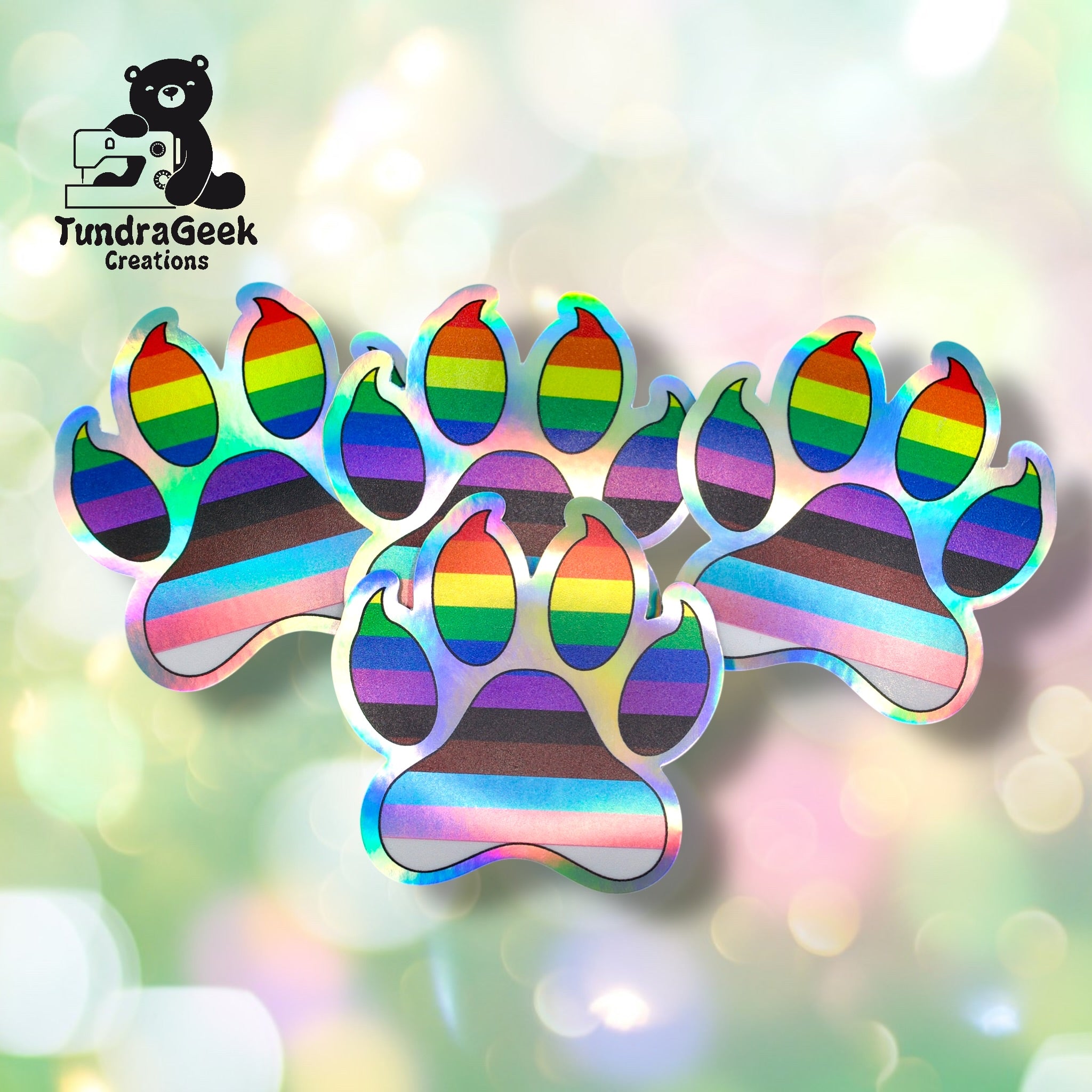 Three holographic paw print stickers with rainbow stripes are displayed on a light, bokeh background. A logo reading TundraGeek Creations appears in the top left corner.