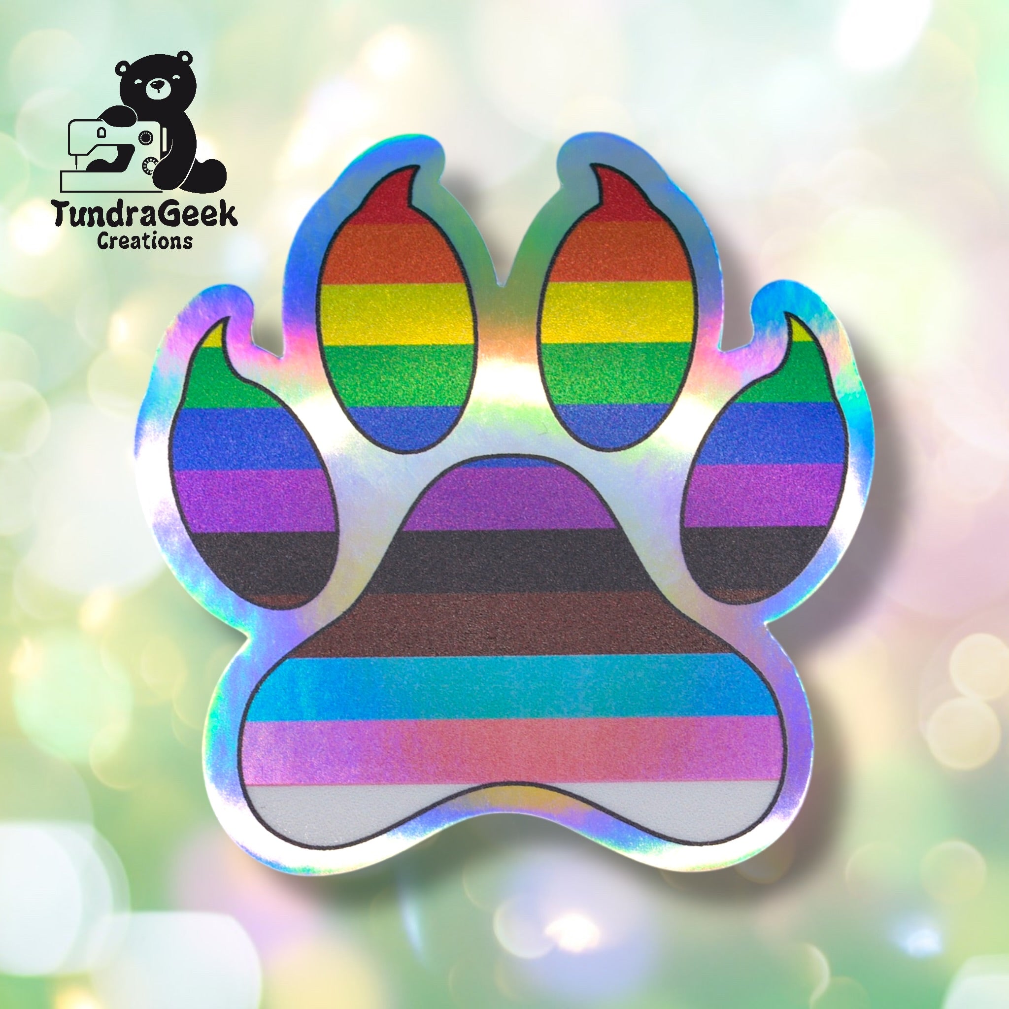 A holographic paw print sticker with rainbow pride stripes, logo in upper left reads TundraGeek Creations with a bear holding a sewing machine.