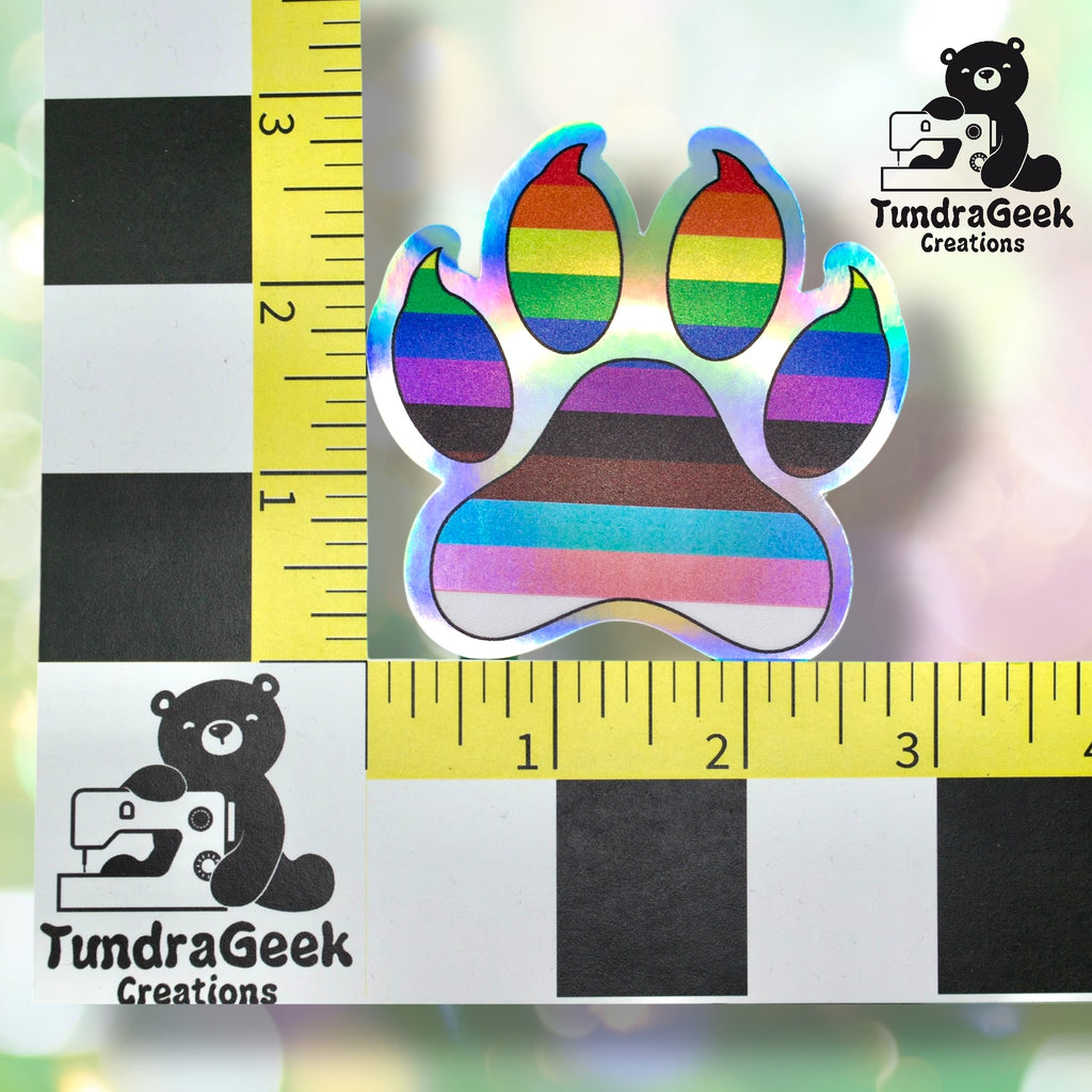 A holographic paw print sticker with rainbow stripes is placed on a measurement grid, showing it is about 2 inches wide. TundraGeek Creations logo appears in two corners.