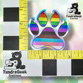 A holographic paw print sticker with rainbow stripes is placed on a measurement grid, showing it is about 2 inches wide. TundraGeek Creations logo appears in two corners.