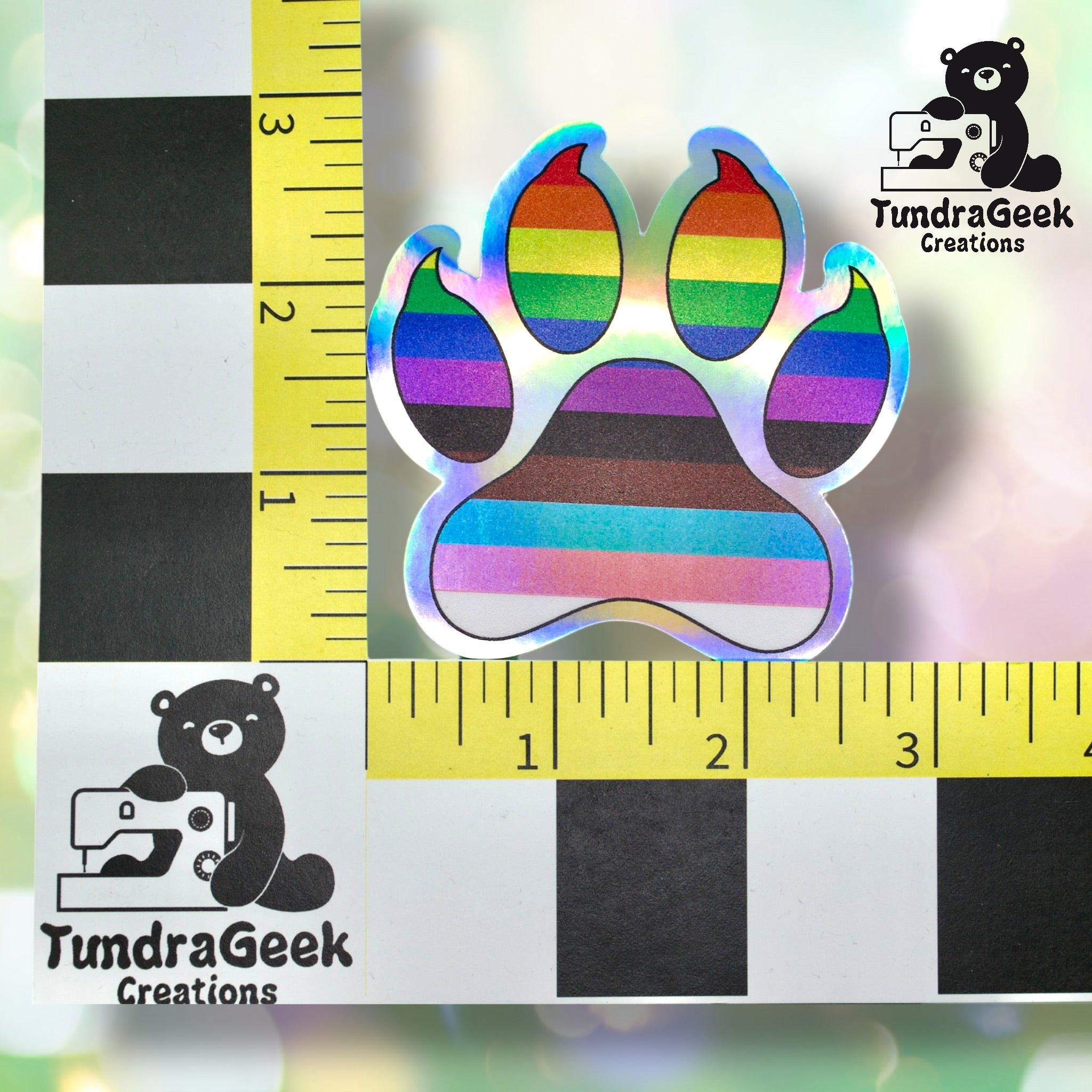 A holographic paw print sticker with rainbow stripes is placed on a measurement grid, showing it is about 2 inches wide. TundraGeek Creations logo appears in two corners.