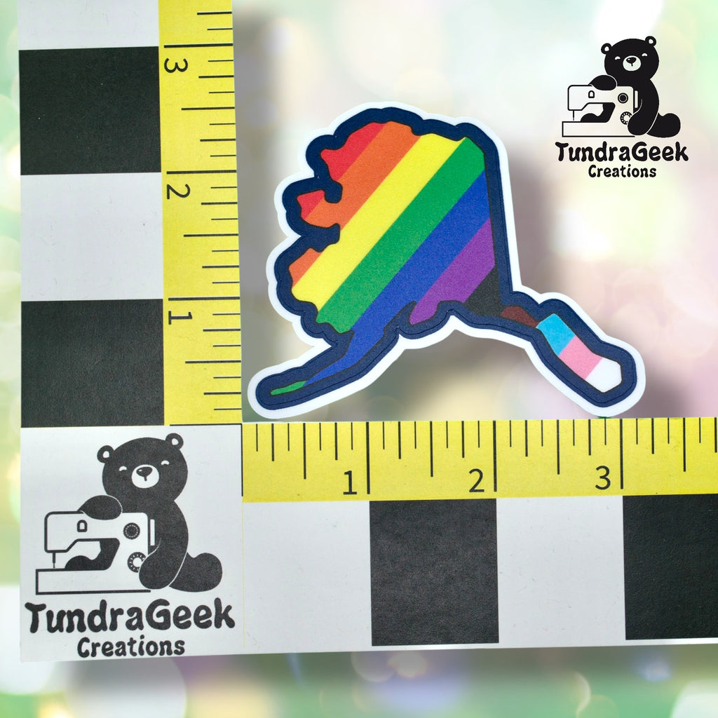 A sticker in the shape of Alaska with a rainbow stripe design is shown next to a ruler and a TundraGeek Creations logo featuring a bear with a sewing machine.