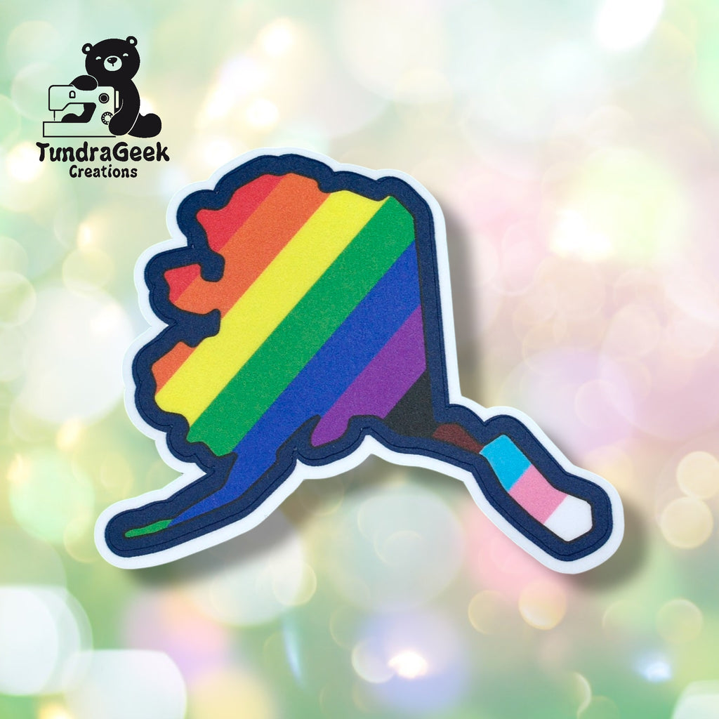 A sticker shaped like Alaska features rainbow stripes and a pink, blue, and white section. The background is blurred with light spots. TundraGeek Creations logo appears in the corner.
