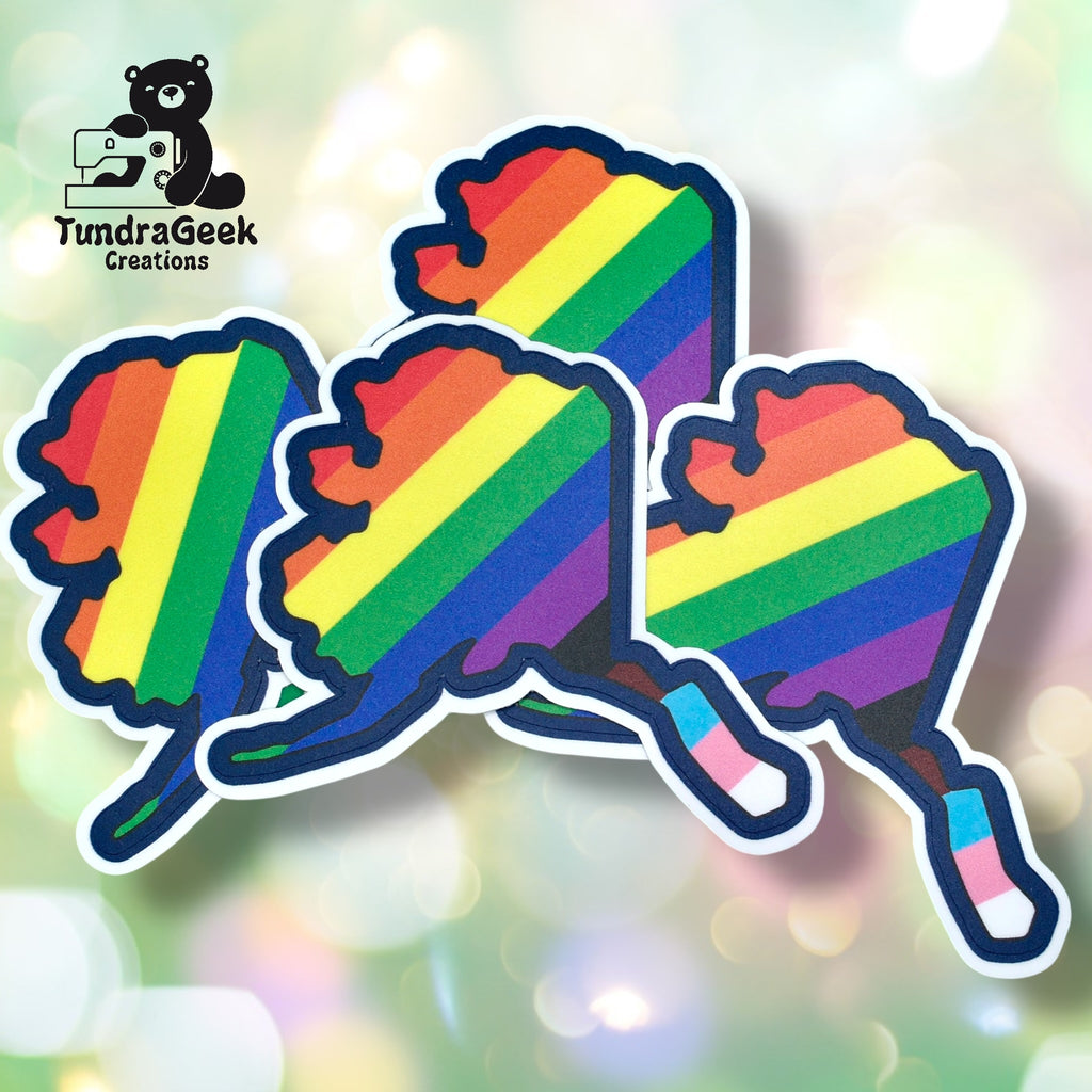 Four stickers of Alaskas outline are filled with rainbow stripes, with the states southeast area colored as a transgender pride flag. TundraGeek Creations logo is in the top left corner.