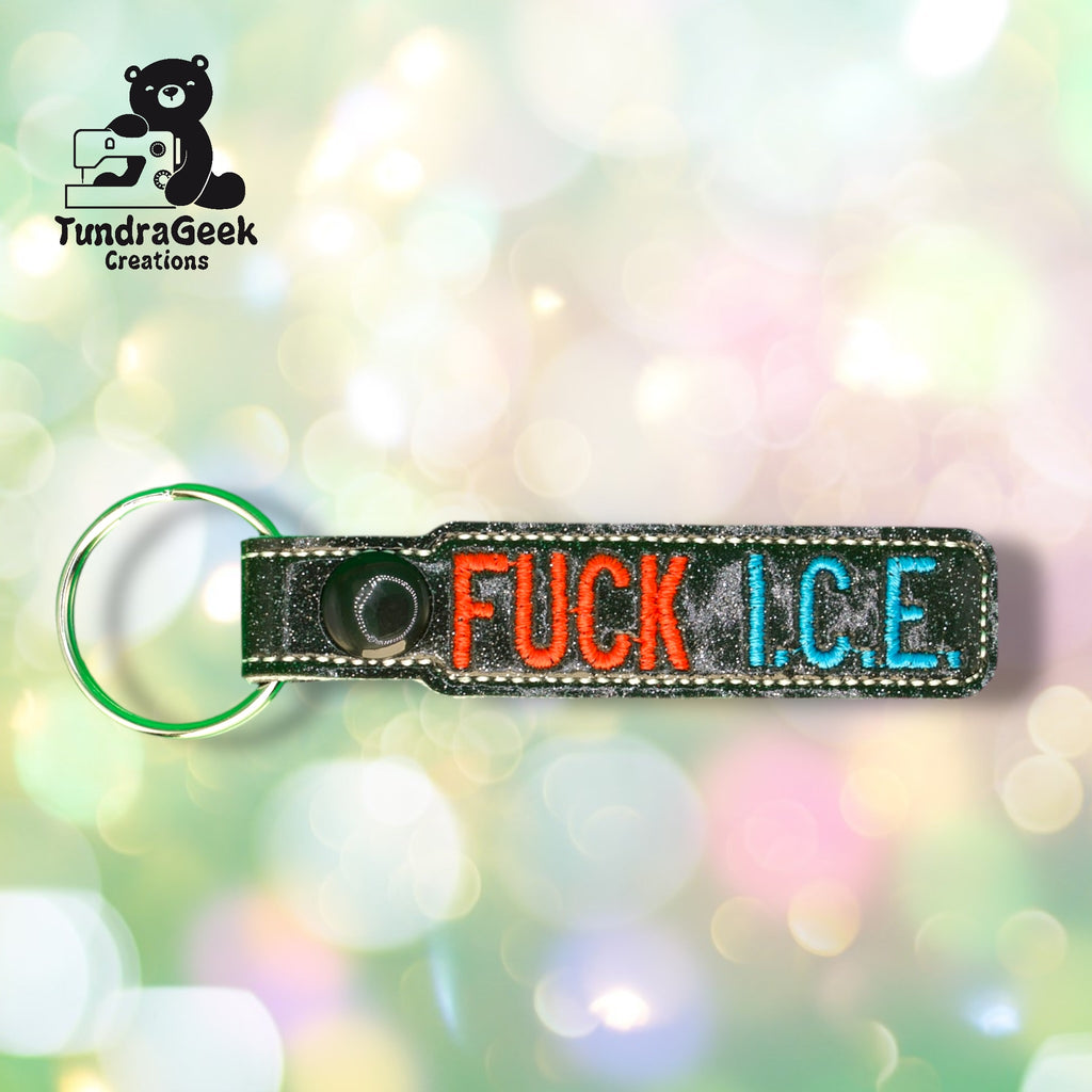 A keychain with the words FUCK I.C.E. embroidered in red and blue letters, with a metal ring, is shown against a blurred, colorful background.