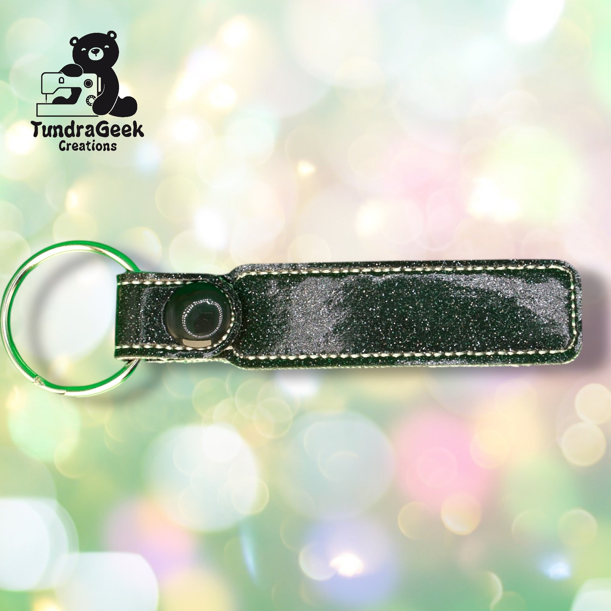 A black glitter keychain holder with white stitching is displayed on a pastel bokeh background. The TundraGeek Creations logo is in the top left corner.