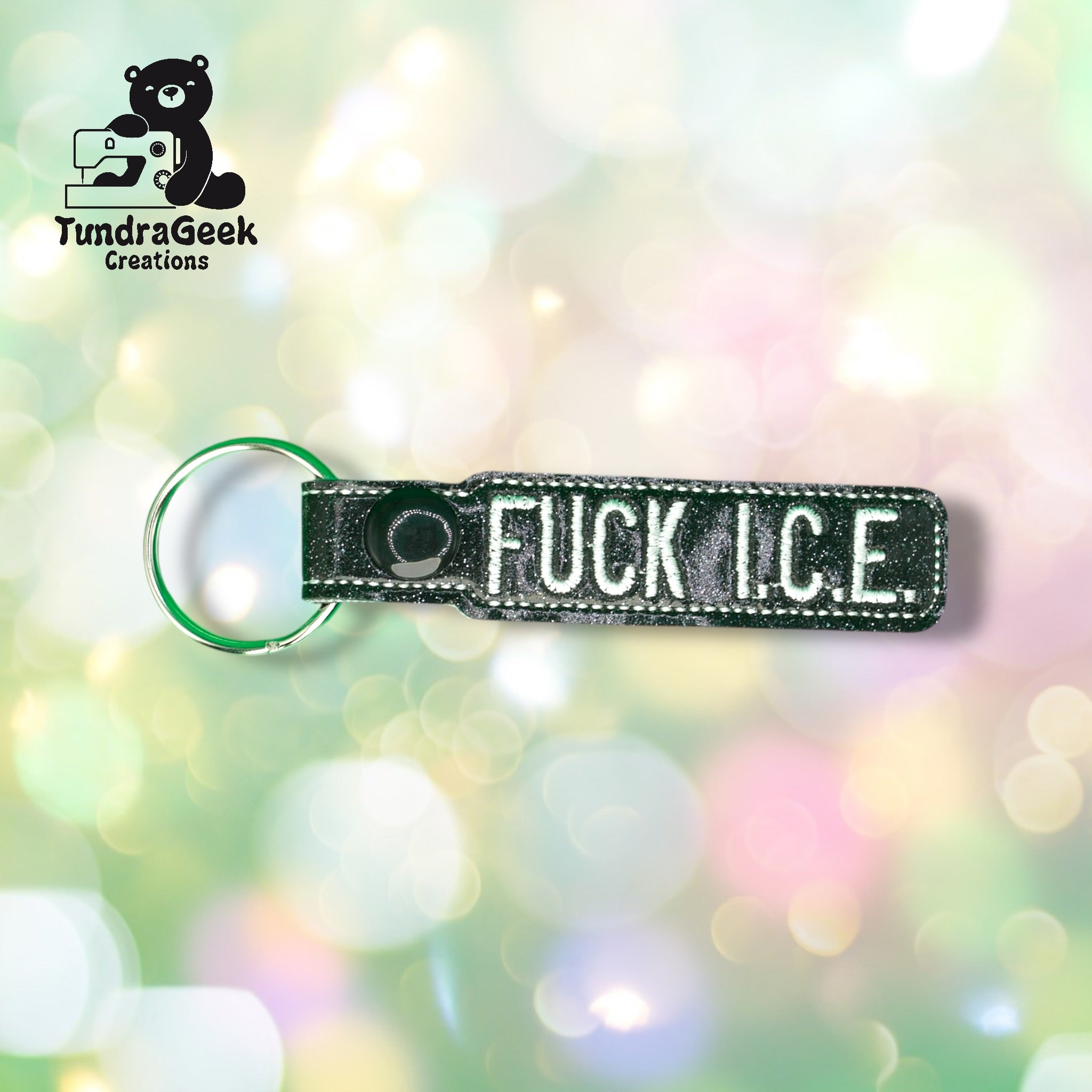 A black keychain with white embroidered text reading FUCK I.C.E. is shown on a blurred pastel background. TundraGeek Creations logo is in the top left corner.