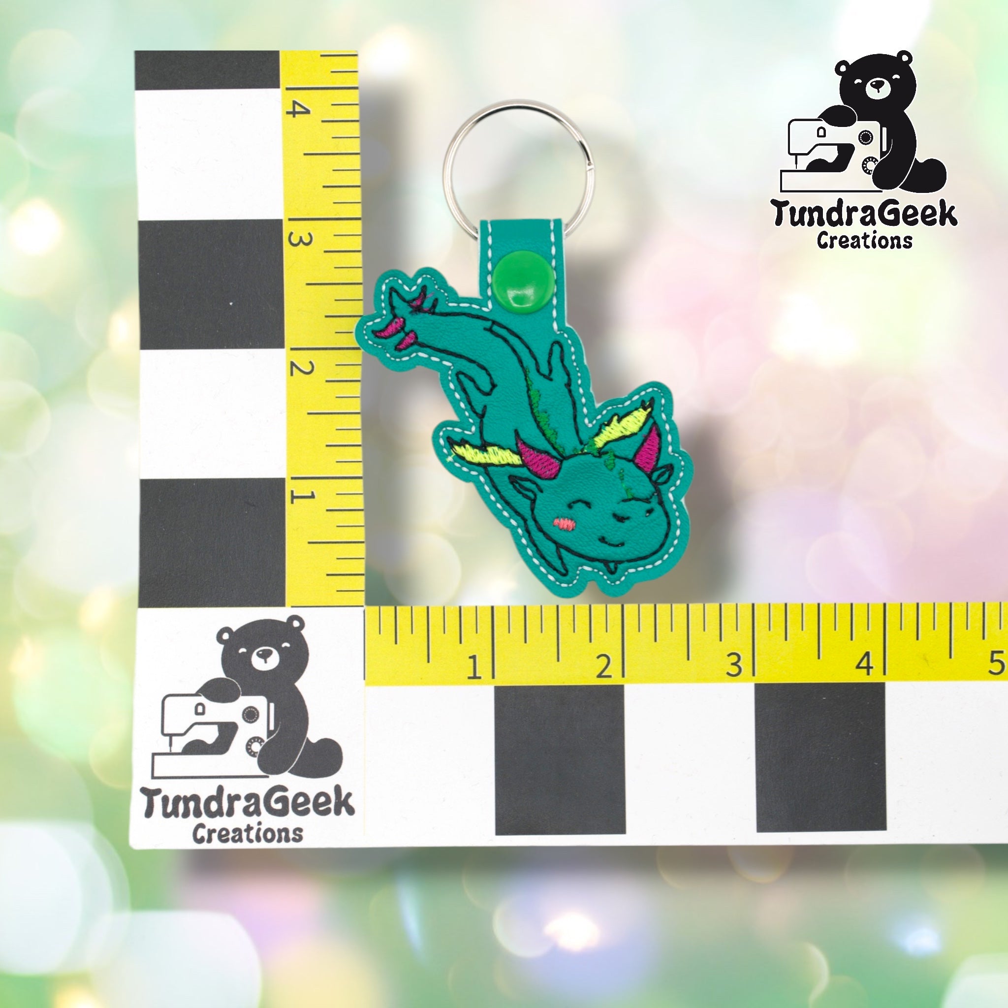 Whimsical dragon keychain positioned against a TundraGeek Creations branded ruler card showing yellow tape measure. Keychain body measures approximately 2.5 inches wide by 3.625 inches tall, not including hardware.