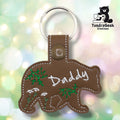 Brown faux leather keychain shaped like a walking bear. White script embroidery reads “Daddy” across the body; green botanical and wildflower embroidery fills the remaining space. Olive plastic snap closure and silver key ring at top. TundraGeek Creations logo in upper right corner.
