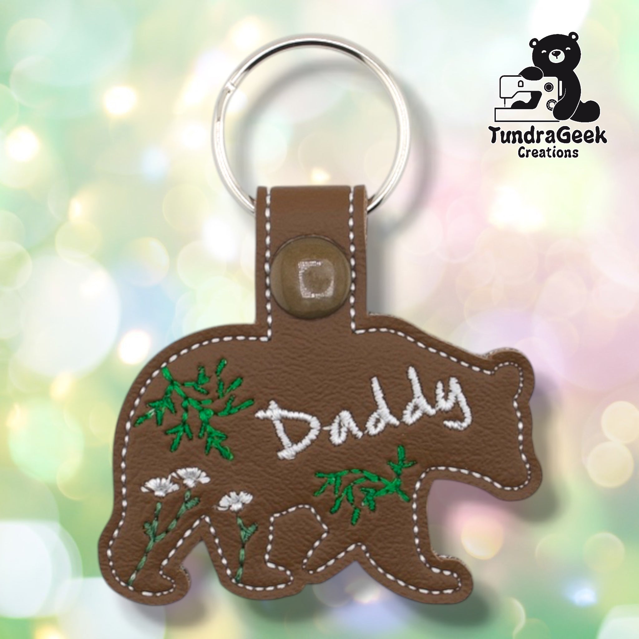 Brown faux leather keychain shaped like a walking bear. White script embroidery reads “Daddy” across the body; green botanical and wildflower embroidery fills the remaining space. Olive plastic snap closure and silver key ring at top. TundraGeek Creations logo in upper right corner.