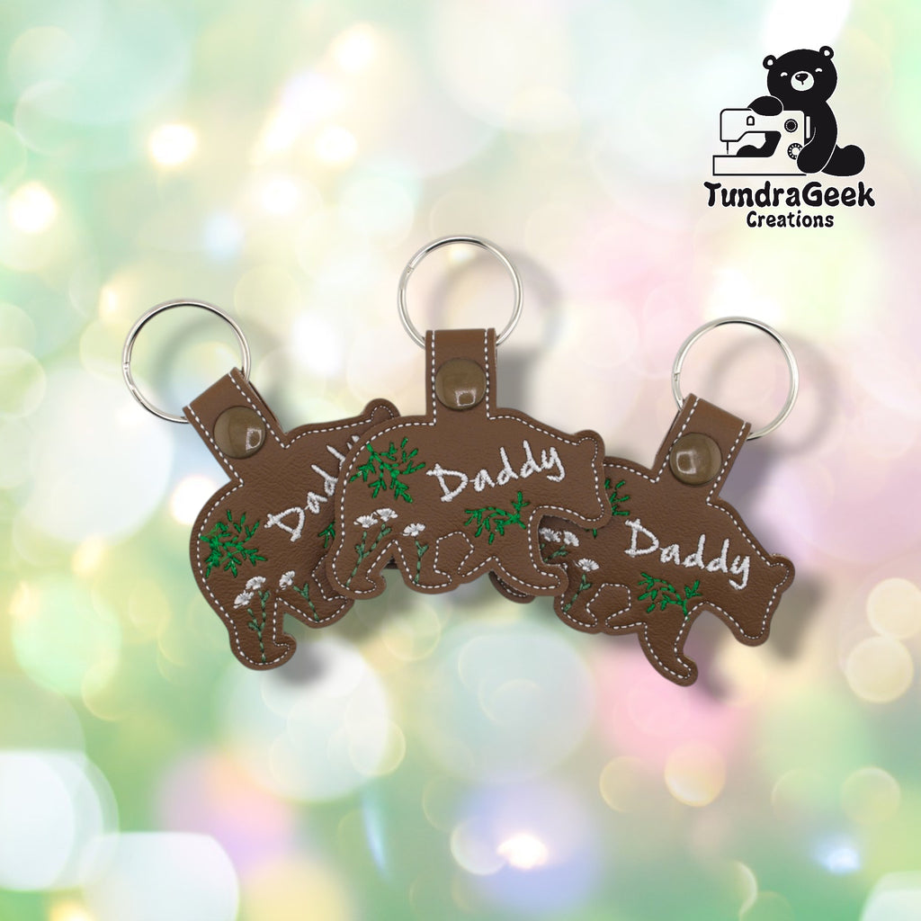 Three Daddy Bear keychains arranged in a row on a pastel bokeh background, showing the front design from slightly varied angles. All three feature matching brown faux leather, white “Daddy” script, and green botanical embroidery.