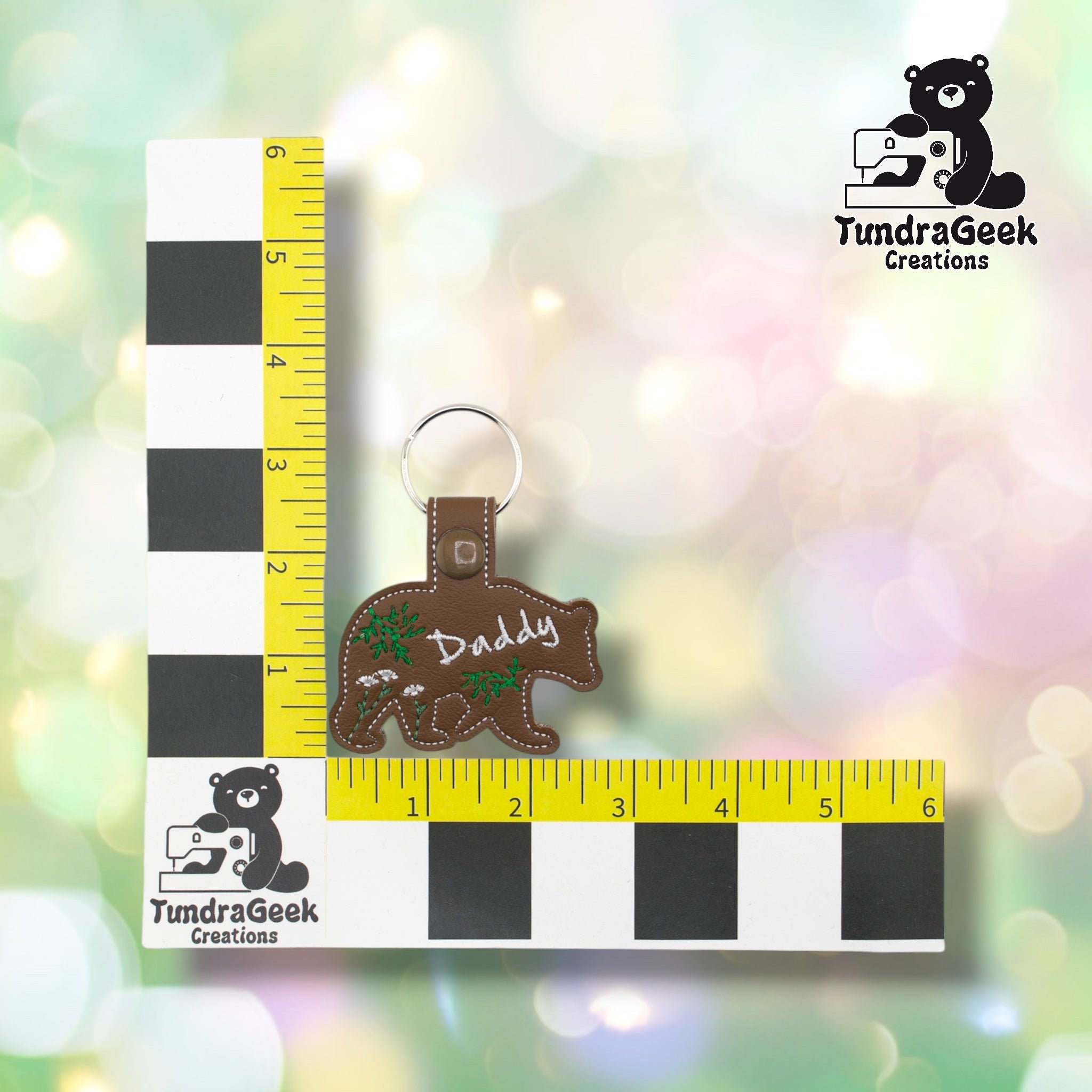 Daddy Bear keychain positioned against a TundraGeek Creations branded ruler card showing yellow tape measure. Keychain body measures approximately 3.5 inches wide by 1.75 inches tall, not including hardware.