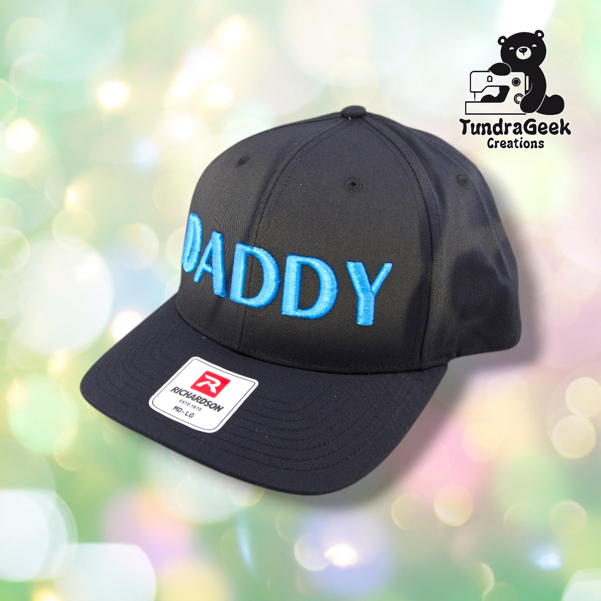 A black Richardson baseball cap with bright blue embroidered "DADDY" text and a white brand label on the front, displayed against a soft pastel bokeh background.