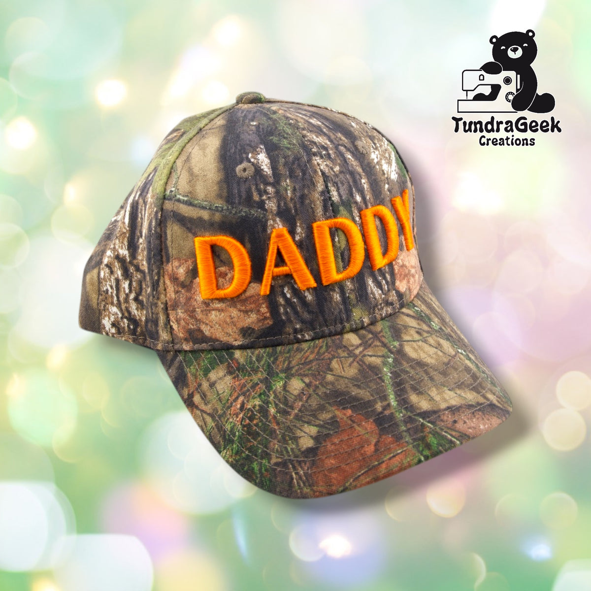 A camouflage baseball cap with orange embroidered text reading "DADDY" against a soft-focused pastel background.