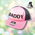A pink and charcoal Richardson trucker cap with "DADDY" embroidered on the front, displayed against a soft blurred green and white bokeh background.
