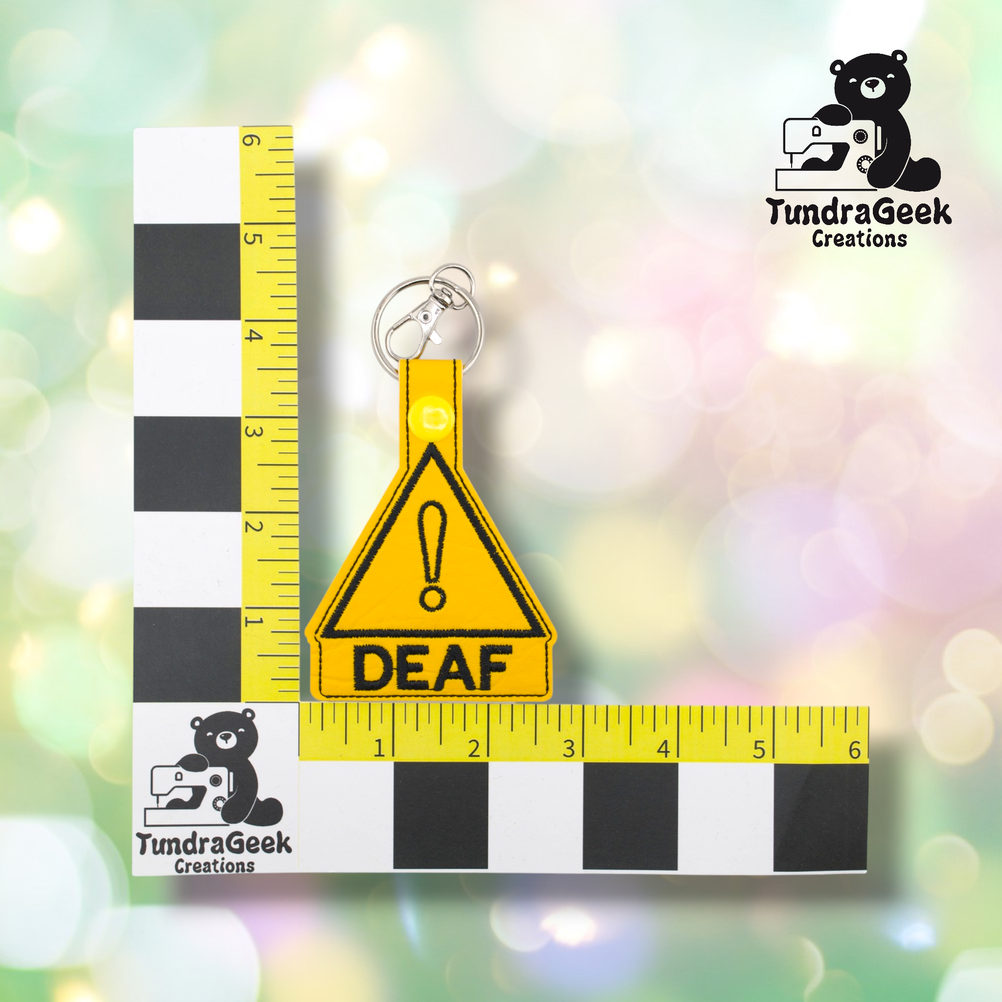 Yellow triangular keychain with a black exclamation mark and the word DEAF, placed on a measurement grid for scale, with the TundraGeek Creations logo in the corners.