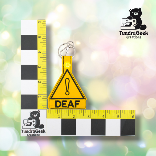 Yellow triangular keychain with a black exclamation mark and the word DEAF, placed on a measurement grid for scale, with the TundraGeek Creations logo in the corners.