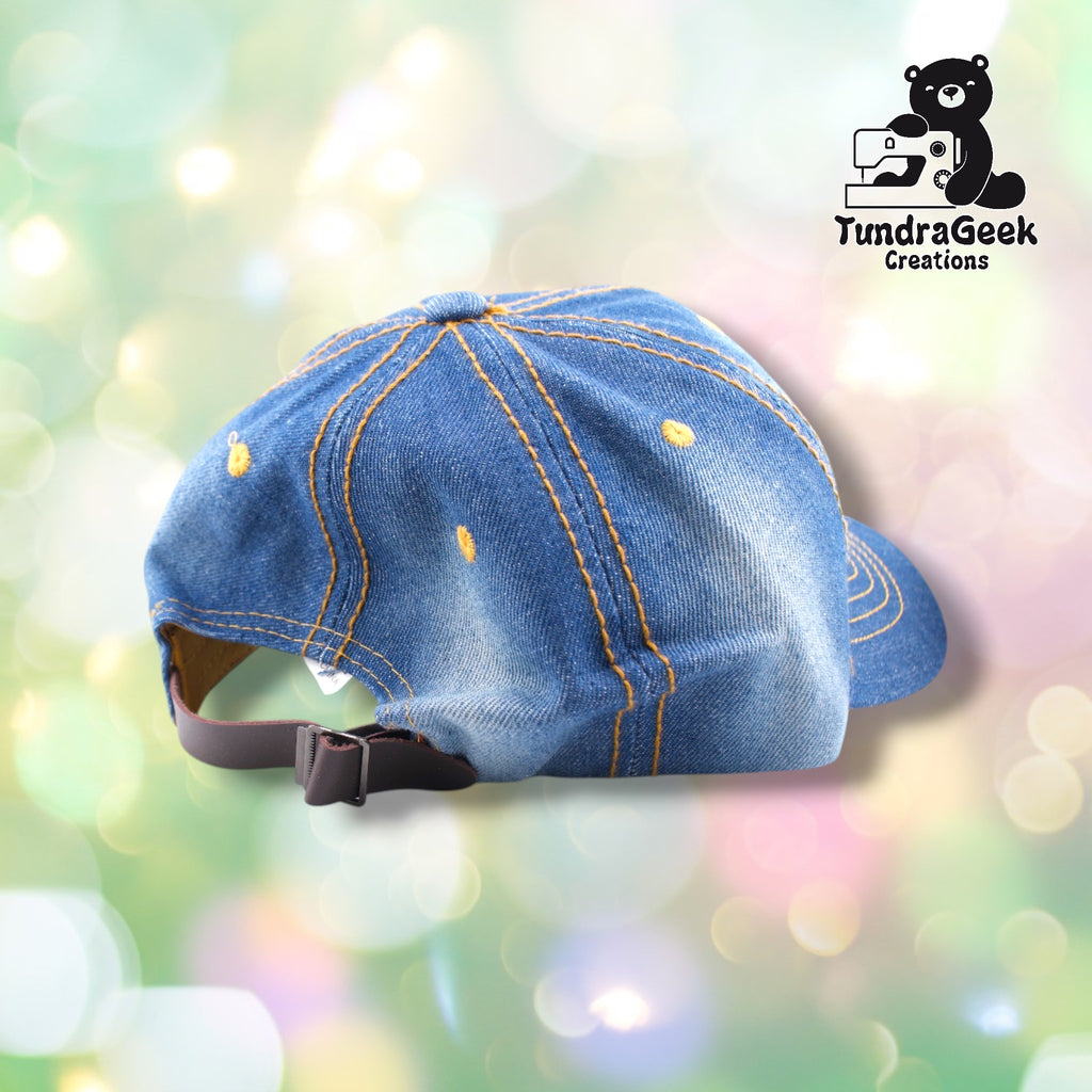 A blue denim baseball cap with orange topstitching displayed on a pastel bokeh background.