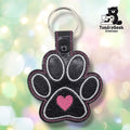 Black glitter faux leather keychain shaped like a dog paw. White embroidery outlines the four toe beans and main pad; a pink heart is embroidered in the center of the main pad. Pink stitching follows the perimeter. Black plastic snap closure and silver key ring at top. TundraGeek Creations logo in upper right corner.