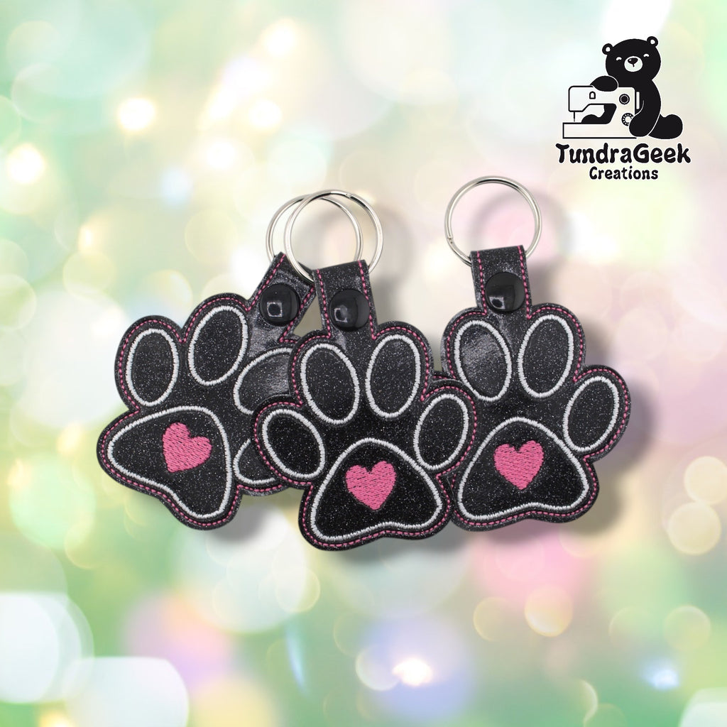 Three dog heart paw keychains arranged together on a pastel bokeh background, showing the design from slightly varied angles. All three feature matching black glitter faux leather, white toe bean embroidery, pink heart centers, and black snap closures.