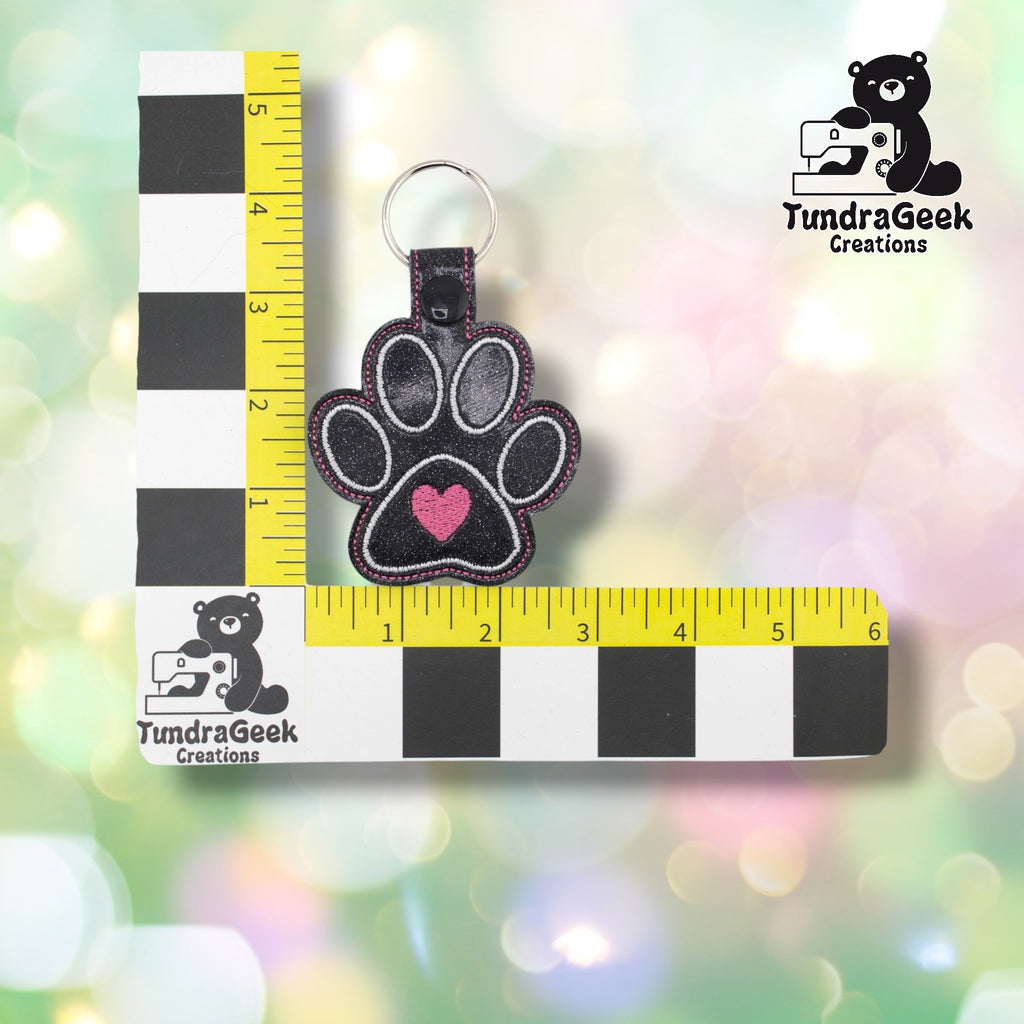 Dog heart paw keychain positioned against a TundraGeek Creations branded ruler card showing yellow tape measure. Keychain body measures approximately 3 inches wide by 2.75 inches tall, not including hardware. Total length with hardware is approximately 4.5 inches.