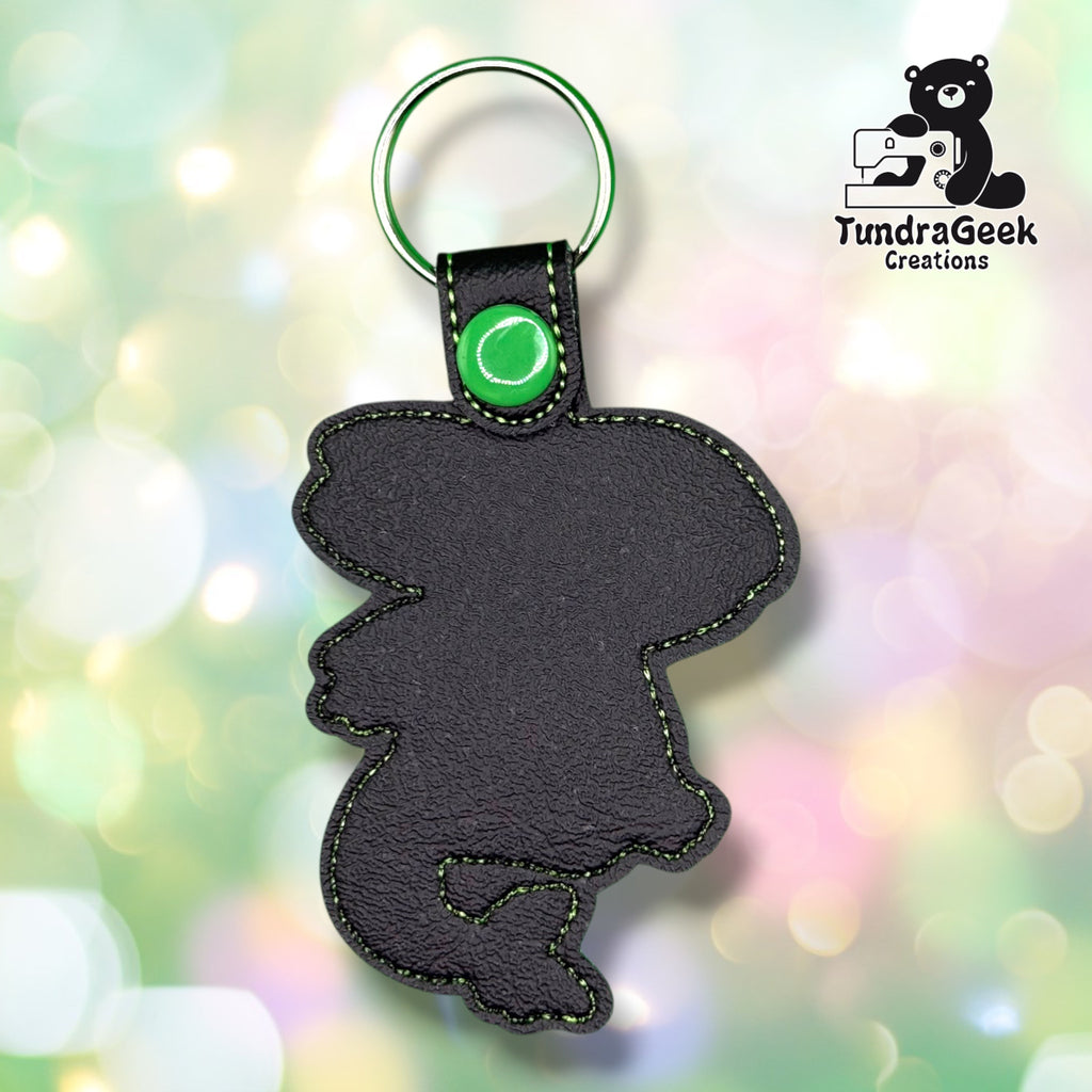 A black faux-leather keychain shaped like a cartoon character, featuring green contrast stitching, a green snap button, and a silver split key ring. The keychain is displayed against a colorful, blurred bokeh background with a 'TundraGeek Creations' logo in the top right corner.