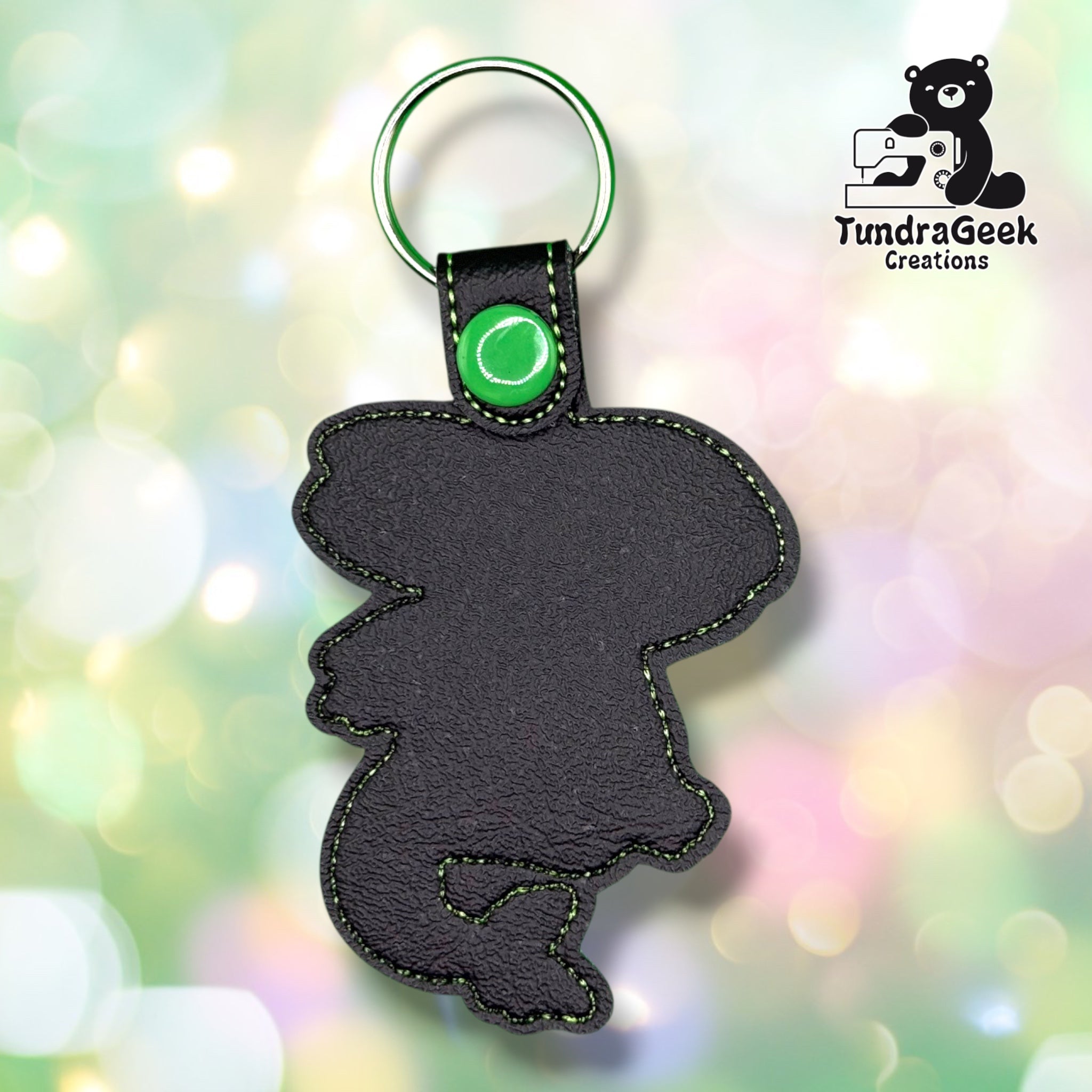 A black faux-leather keychain shaped like a cartoon character, featuring green contrast stitching, a green snap button, and a silver split key ring. The keychain is displayed against a colorful, blurred bokeh background with a 'TundraGeek Creations' logo in the top right corner.