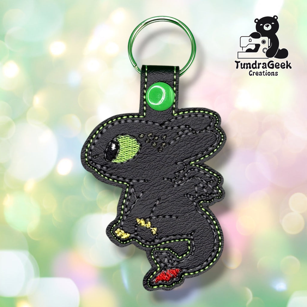 A black faux leather patch keychain shaped like a cartoon dragon with green stitching, a silver ring, and small red and yellow accents, shown against a soft pastel bokeh background. The TundraGeek Creations logo is visible in the top right.