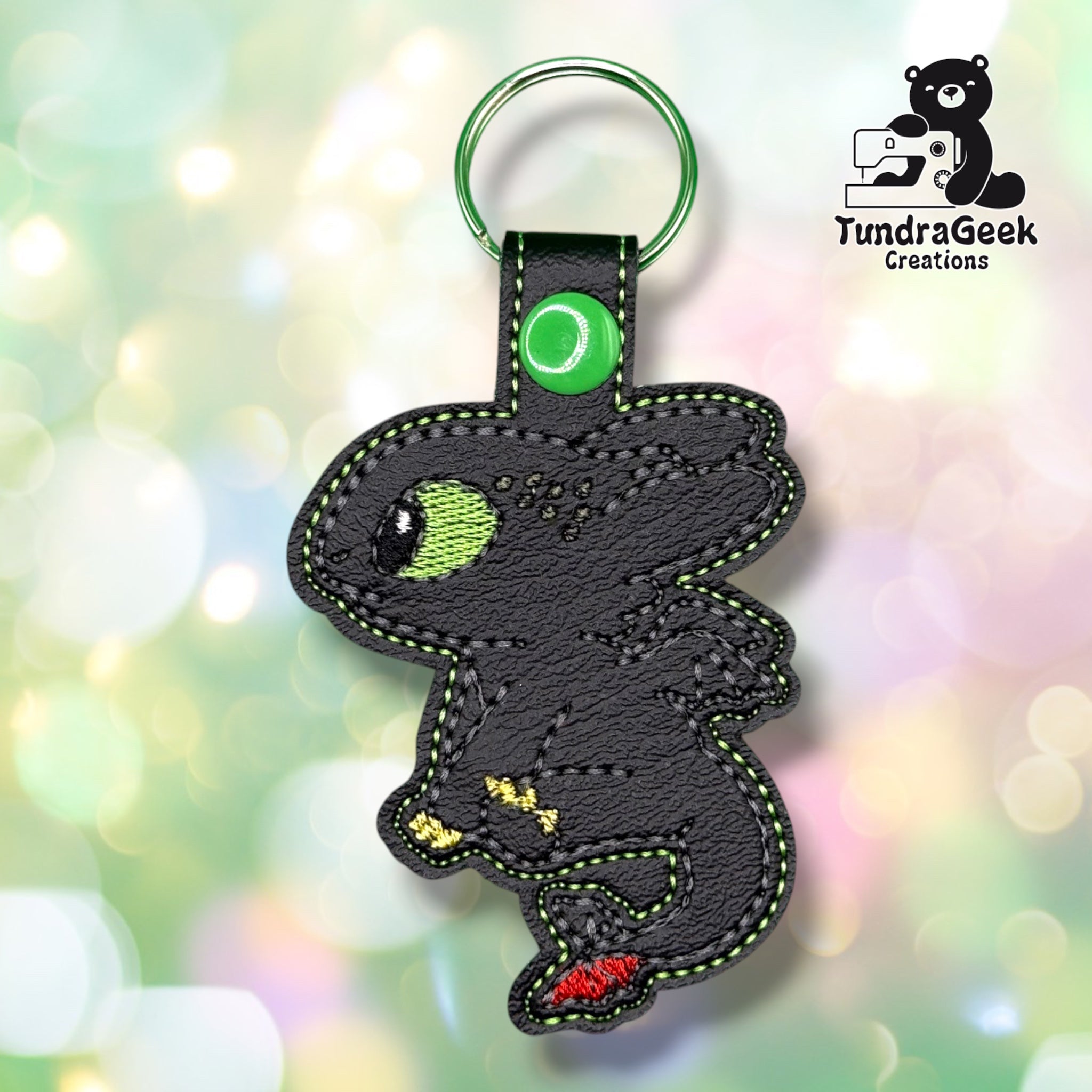 A black faux leather patch keychain shaped like a cartoon dragon with green stitching, a silver ring, and small red and yellow accents, shown against a soft pastel bokeh background. The TundraGeek Creations logo is visible in the top right.