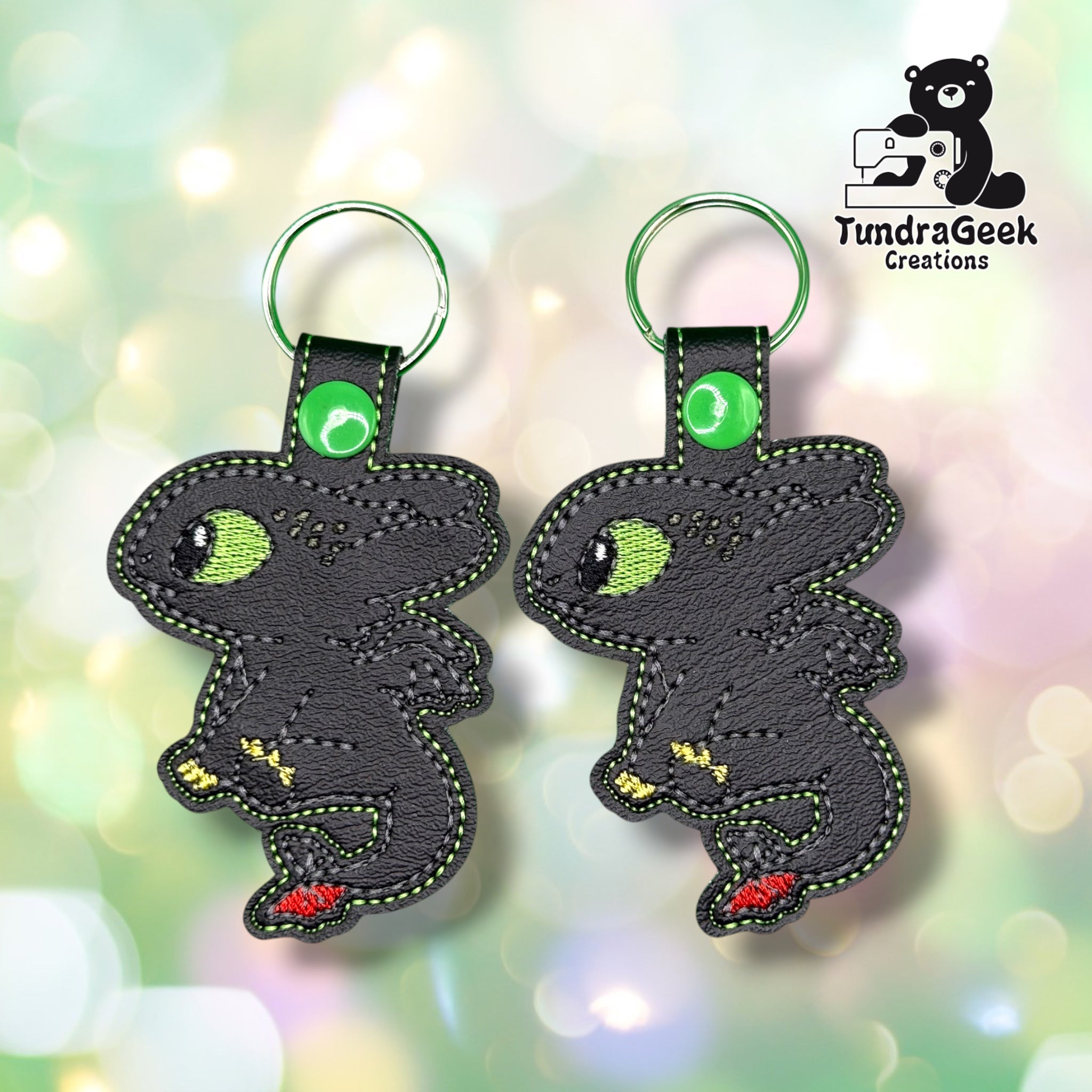 Two black dragon-shaped keychains with green stitching, green eyes, and small red and yellow accents, shown on a soft pastel bokeh background. A TundraGeek Creations logo appears in the top right corner.