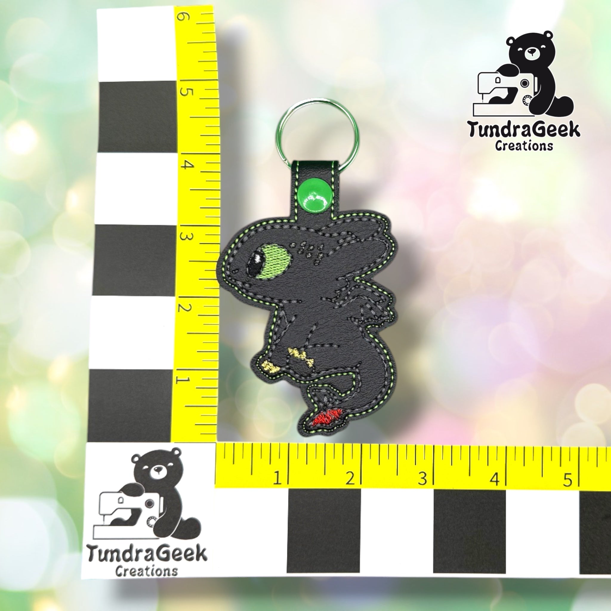 A small embroidered dragon keychain shown upright beside a black-and-white checkerboard ruler and yellow measuring tape, with the TundraGeek Creations logo in the top right and bottom left.