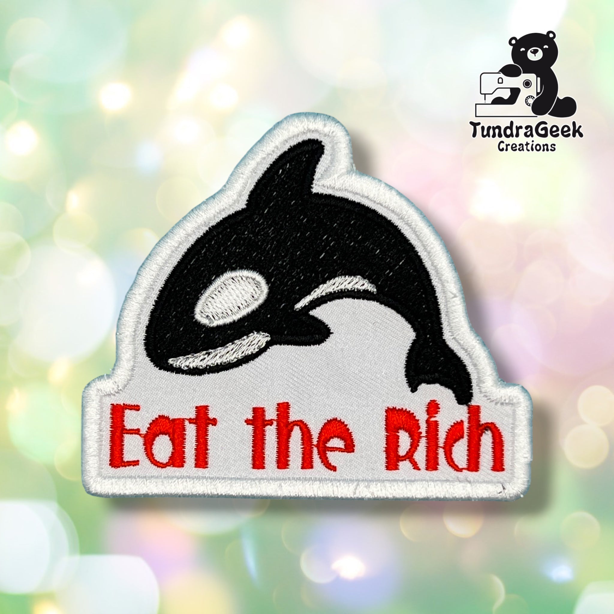 An embroidered patch featuring a black and white orca above the red text 'Eat the Rich' on a white background, shown against a soft pastel bokeh backdrop. A TundraGeek Creations logo is visible in the top right corner.