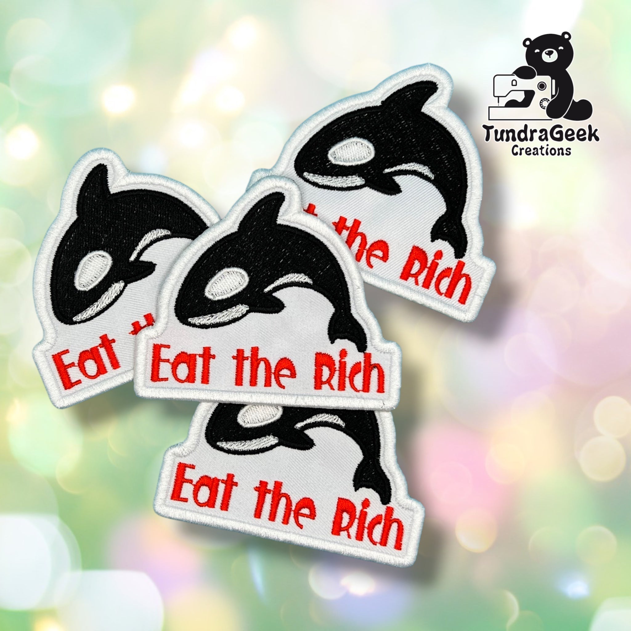 Four embroidered iron-on patches featuring a black and white orca fish design with the red text 'Eat the Rich' on a white background, arranged on a colorful bokeh backdrop. A TundraGeek Creations logo is visible in the top right corner.
