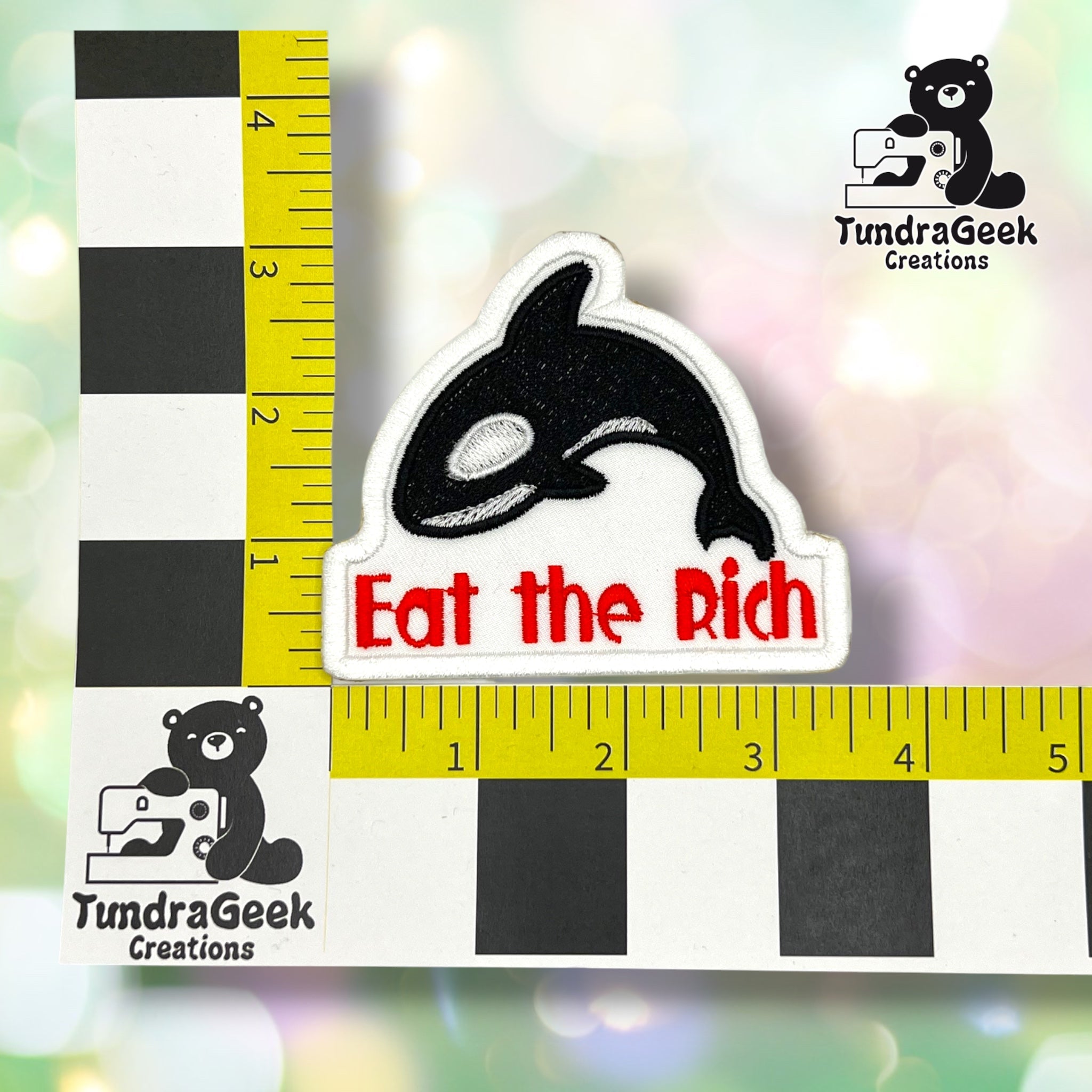 A die-cut embroidered patch featuring a black and white orca fish design above the red text 'Eat the Rich' on a white background. The patch is displayed with a yellow-and-black measurement ruler for scale, with the TundraGeek Creations logo visible in the top-right and bottom-left corners.