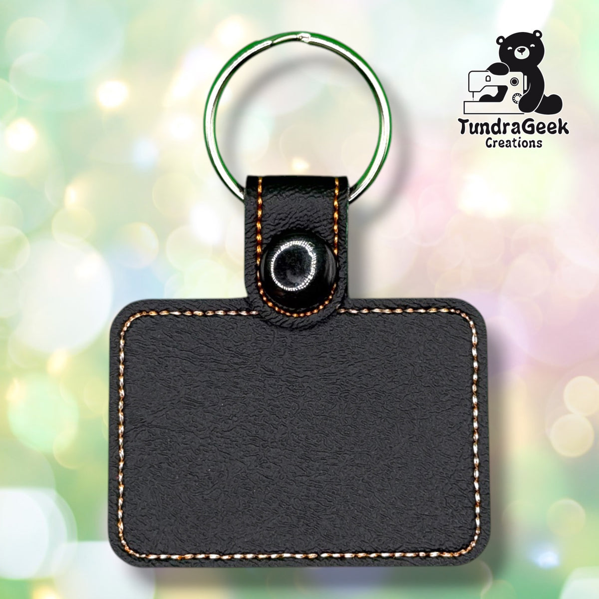 Back of a black vinyl keychain showing the plain backing with tan and white stitched outline, a black plastic snap closure, and a green split ring.