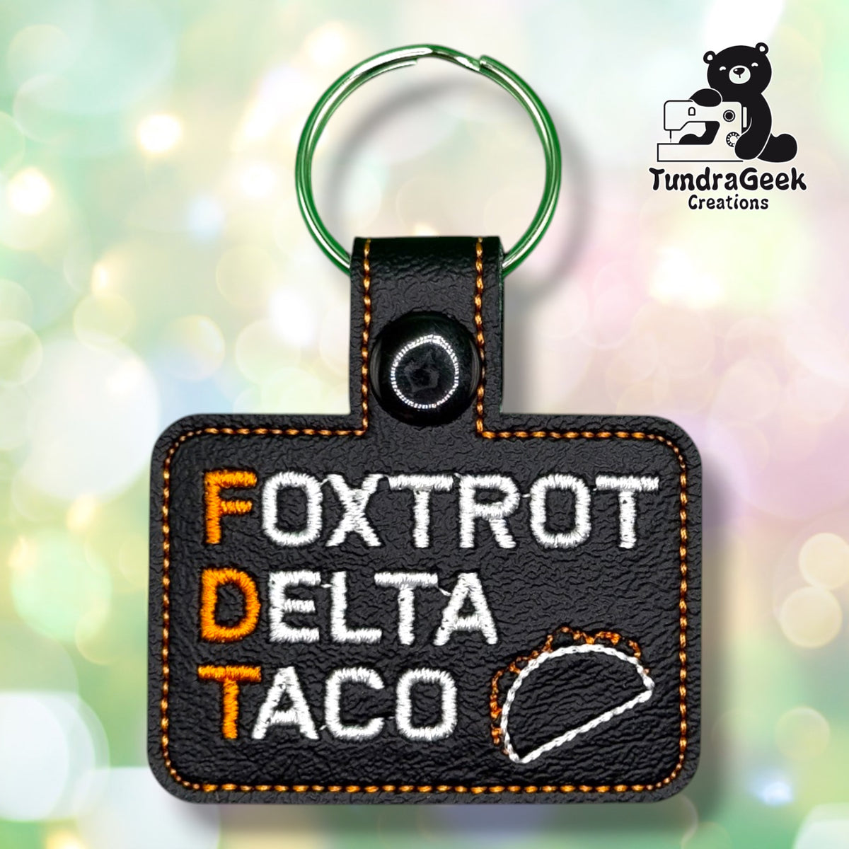 Front view of a black vinyl keychain reading "FOXTROT" and "DELTA" in white with orange first letters, and "TACO" in white with an orange T, next to a stitched taco outline. Finished with a black plastic snap closure and green split ring.