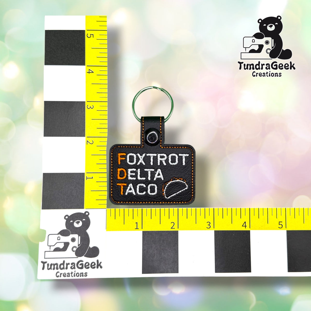 Keychain shown against a black-and-white checkered background with yellow rulers for scale, measuring approximately 2.5 inches wide by 3.5 inches tall, including hardware.  ---