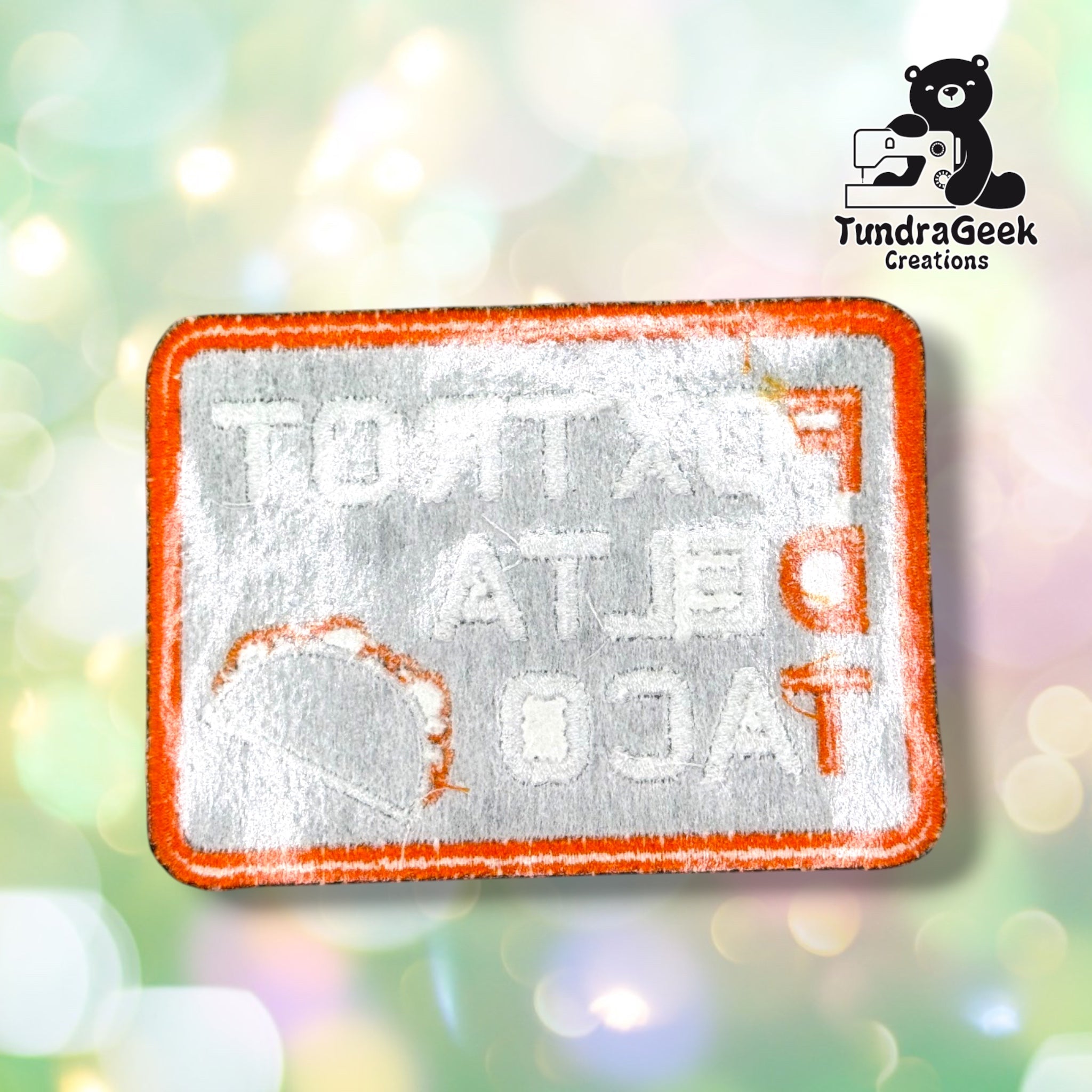 Back of the patch showing the reverse side of the embroidery in muted grey and orange thread, with the lettering and taco outline mirrored and visible through the backing, and the orange merrowed edge around the perimeter.