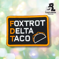 Front view of a black rectangular patch with an orange merrowed edge, reading "FOXTROT", "DELTA", and "TACO" in white with orange first letters, next to a white-and-orange stitched taco outline.