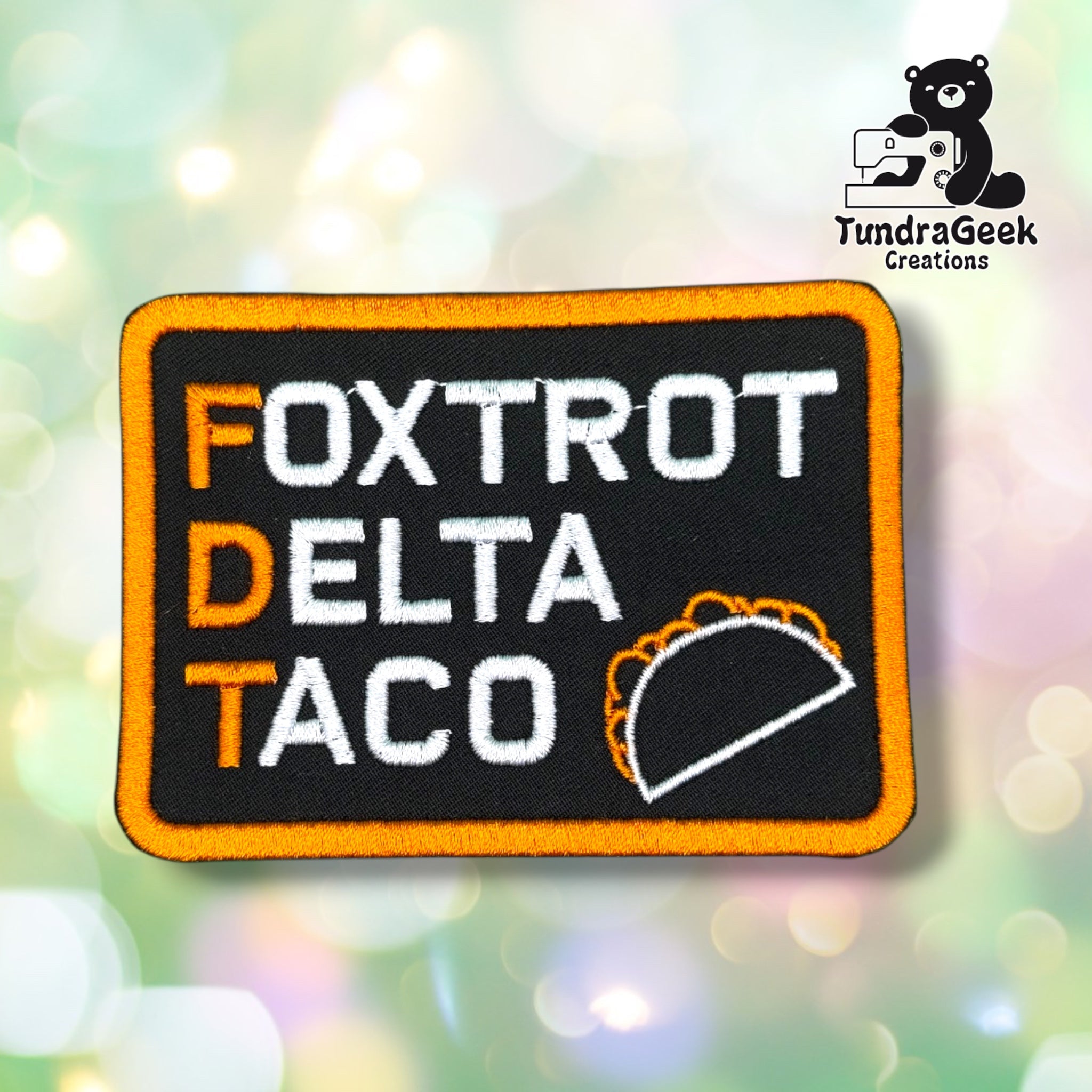 Front view of a black rectangular patch with an orange merrowed edge, reading "FOXTROT", "DELTA", and "TACO" in white with orange first letters, next to a white-and-orange stitched taco outline.