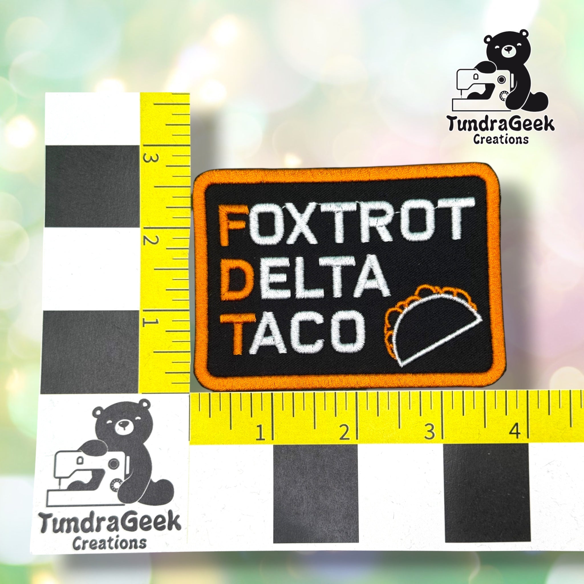 Patch shown against a black-and-white checkered background with yellow rulers for scale, measuring approximately 3.75 inches wide by 2.75 inches tall.  ---